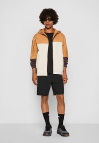 Sudadera con cremallera bloqueada por colores en marrón, crema y mangas oscuras; camiseta negra; pantalones cortos negros; zapatillas negras; gafas de sol redondas.