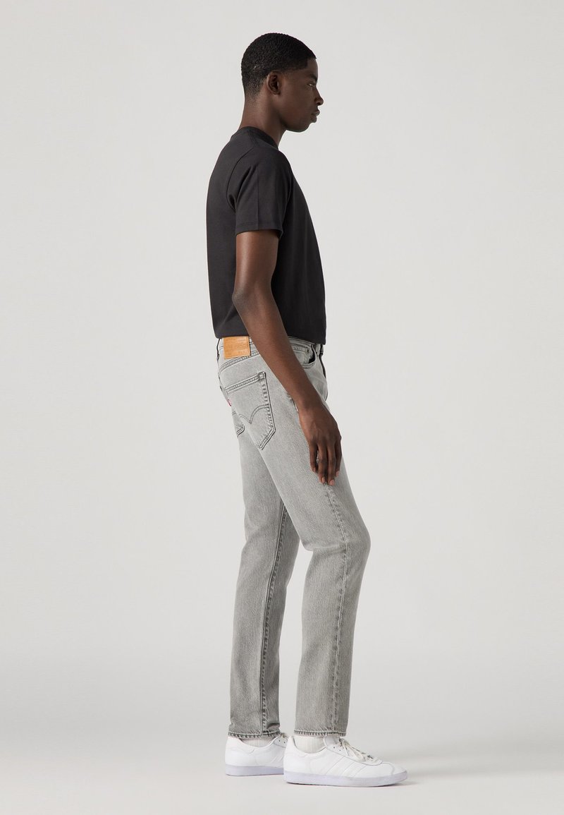 Grijze jeans met een rechte snit en een leren patch op de achterkant. Gepaard met een zwart T-shirt en witte sneakers. Eenvoudig ontwerp, casual stijl.