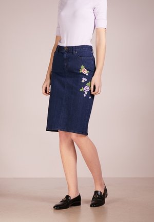 Vrouw draagt een donkerblauwe denim rok tot op de knieën met bloemenborduursel aan de zijkant en zwarte loafers, gecombineerd met een lichtroze top.