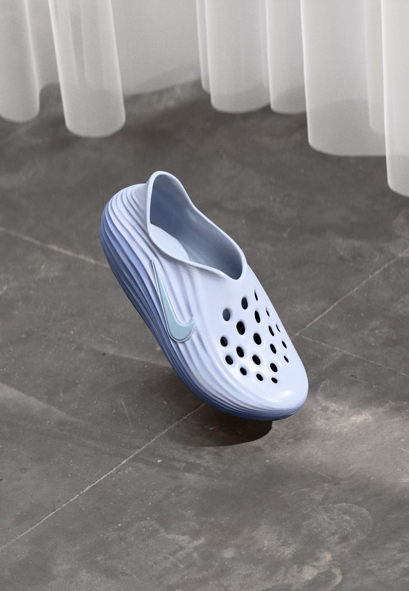 Zapatilla azul clara de Nike, tipo slip-on, con agujeros de ventilación y suela estriada, flotando sobre un suelo de azulejos grises bajo cortinas blancas translúcidas.