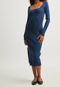Abito lungo a maniche lunghe blu con costine, caratterizzato da una scollatura quadrata, una forma aderente e uno spacco laterale, abbinato a ballerine nere.
