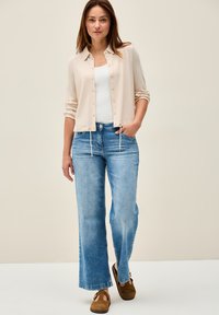 Beige knapp-up kofta med rullade ärmar över en vit linne, tillsammans med ljusblå vida jeans och bruna mockaskor.