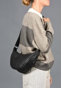 Bolso de mensajero de cuero negro con forma curva, con una textura suave y herrajes plateados. Lleva puesto sobre un suéter de rayas beige y gris.