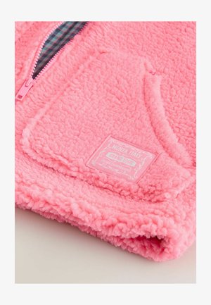 Rožinės spalvos fliso striukė su tekstūruota, pūkuota danga. Yra užtrauktukas, kišenė ant pilvo ir audinio žymė su užrašu „Monza, Italija“.