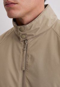 Beżowa kurtka z wysokim kołnierzem, zapinana na zamek i dwa zatrzaski. Miękki, gładki materiał o subtelnym połysku.