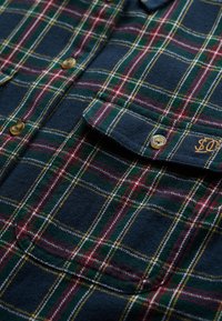 Chemise en flanelle avec une base bleu marine et un motif à carreaux multicolore en vert, rouge et jaune. Inclut une poche arrondie et des boutons en laiton.