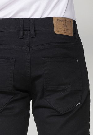 Pantaloni in denim neri con una toppa in pelle in vita, dotati di due tasche posteriori con pattine angolate e un sottile dettaglio di cucitura sul tessuto.