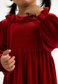 Czerwona sukienka z aksamitnym wykończeniem, z bufkami i falbaniastym dekoltem. Posiada plisowaną spódnicę i miękką fakturę. W kadrze częściowo widoczna ręka.