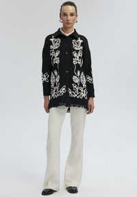 Cardigan lungo nero con ricami floreali bianchi, chiusura con bottoni e orlo frangiato, indossato sopra pantaloni bianchi a zampa d'elefante e scarpe nere.