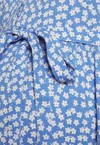 Tela azul con estampado floral blanco, con detalle de cintura anudada y acabado texturizado. El patrón consiste en pequeñas flores dispersas.