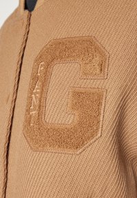 Brązowy sweter dzianinowy z teksturowaną naszywką w kształcie litery "G" oraz wypukłym napisem "GANT". Posiada zamek błyskawiczny i prążkowane wykończenia.