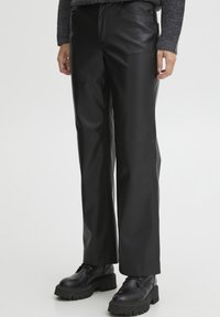 Pantaloni in ecopelle neri con un design a gamba dritta, caratterizzati da cuciture in evidenza e una texture liscia. Indossati con stivali neri chunky.