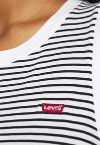 Gros plan sur une personne portant une chemise blanche avec des rayures horizontales noires et un petit écusson rouge avec le logo Levi's sur la poitrine.