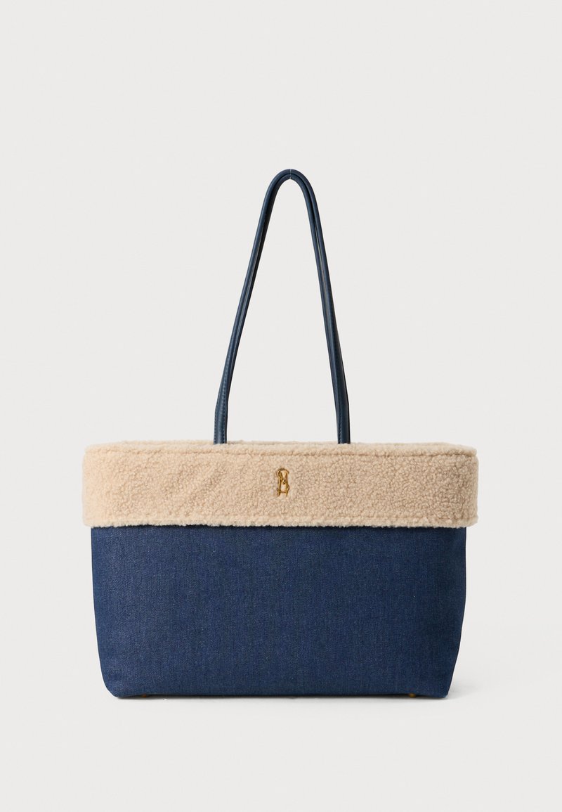 Geantă de umăr din denim cu o margine din blană faux bej, are două mânere și un accent cu logo auriu. Formă rectangulară, material texturat.