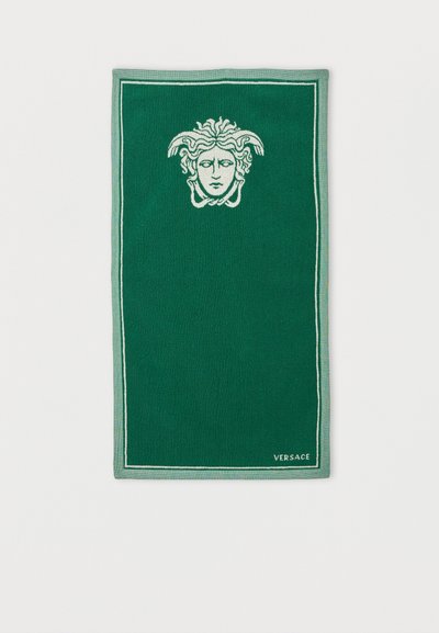 Πράσινη ορθογώνια πετσέτα με λευκό έμβλημα κεφαλής Μέδουσας στο πάνω κέντρο και την επωνυμία «Versace» στην κάτω δεξιά γωνία.