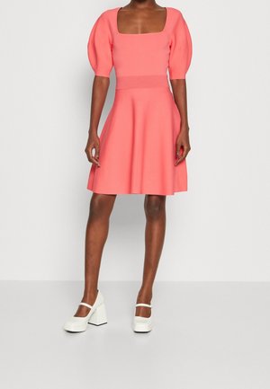 Femme portant une robe corail rose mi-longue avec manches bouffantes et encolure carrée, associée à des chaussures Mary Jane blanches à talon bloc.