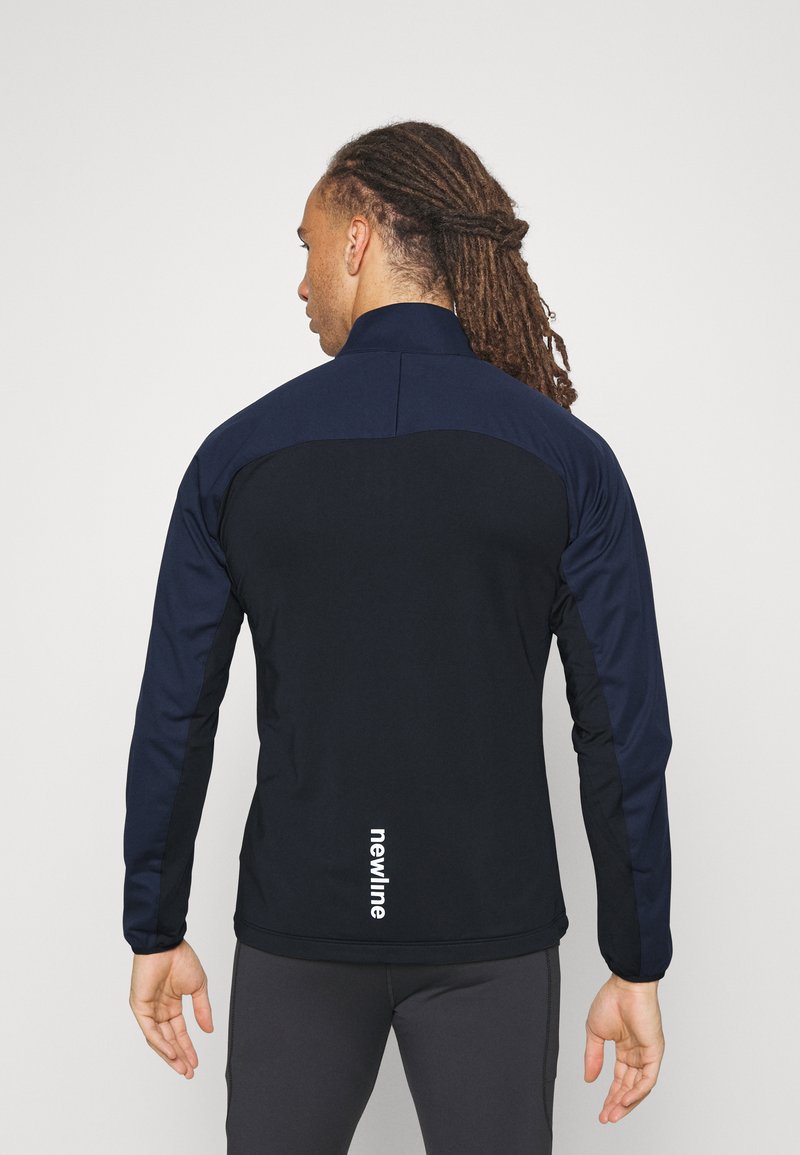 Marineblaue Sportjacke mit hohem Kragen, kontrastierenden Texturen und einem weißen Logo am hinteren Saum. Taillierter Schnitt und lange Ärmel.