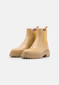 Bottines beige brillantes avec semelles épaisses et panneaux latéraux en elastique sur un fond blanc.