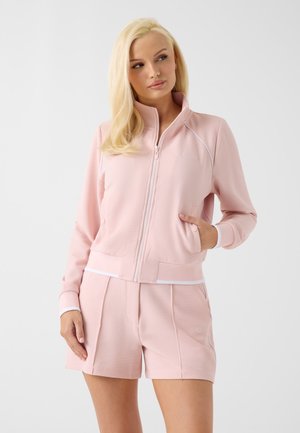 Femme aux longs cheveux blonds portant une veste à fermeture éclair rose clair texturée et un short assorti, debout, une main dans la poche, sur fond blanc.