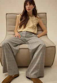 Beige bestickte Bluse kombiniert mit hüftig sitzenden, weit geschnittenen Karohosen in Braun, Blau und Creme, getragen mit braunen Wildlederschuhen.