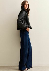 Giacca cropped in ecopelle nera con bordo in pelliccia, abbinata a jeans in denim scuro a vita alta e gamba larga e sandali open-toe.