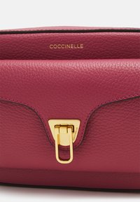 Borsa a tracolla in pelle fucsia con finitura testurizzata, dotata di una chiusura tono oro e del nome del marchio "Coccinelle" impressa in oro.