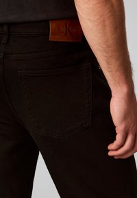 Κοντινό ενός ατόμου που φοράει μαύρα τζιν με καφέ ταμπελάκι λογότυπου Calvin Klein πάνω από την πίσω τσέπη, δείχνοντας μέρος του μπράτσου και του χεριού.