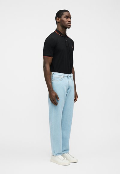 Νέος άνδρας που στέκεται, φοράει μαύρο πόλο με κόκκινο φινίρισμα, ανοιχτόχρωμα τζιν και λευκά αθλητικά παπούτσια, με φόντο μονόχρωμο λευκό.