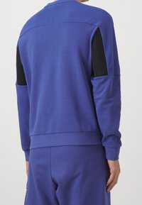 Persona che indossa un top a maniche lunghe blu e pantaloni abbinati con pannelli in rete nera sulle maniche superiori, vista da dietro.