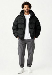 Doudoune noire à capuche avec un motif matelassé et des poignets élastiques, associée à un pantalon cargo gris et des baskets blanches.