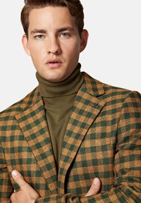 Brązowa i zielona w kratę marynarka o teksturowanym wykończeniu, noszona na ciemnooliwkowym golfie. Marynarka ma klapy z wycięciem i szpilkę.