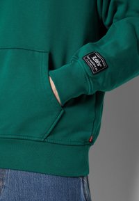 Grüne Fleece-Hoodie mit Kängurutasche, geripptem Saum und Bündchen. Mit einem schwarzen Etikett am Ärmel. Getragen mit blauen Jeans.