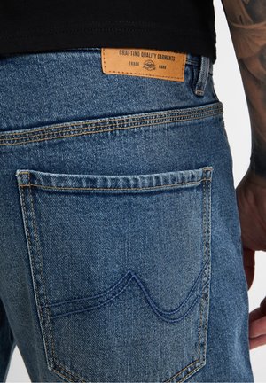 Nahaufnahme einer hinteren Tasche einer blauen Denim-Jeans mit einem Lederetikett mit der Aufschrift „Crafting Quality Garments“, getragen von einer Person mit tätowiertem Arm.