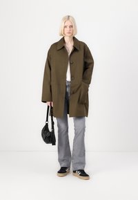 Olivengrønn oversized frakk med krage, knapper foran og sidelommer, kombinert med lysegrå, utsvingte jeans og svarte sko. Svart veske holdt på siden.