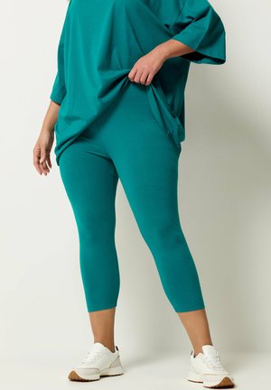 Person trägt türkisfarbene, gekürzte Leggings und passendes übergroßes Oberteil, zieht das Oberteil leicht, mit weißen Turnschuhen vor einfachem Hintergrund.