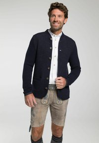 Mann im traditionellen bayerischen Outfit, mit einer marineblauen Strickjacke, weißem Hemd, bestickten Lederhosen und dekorativer Gürtelschnalle, lächelt.