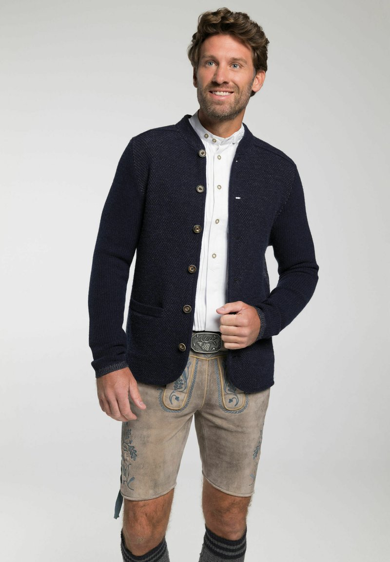 Mann im traditionellen bayerischen Outfit, mit einer marineblauen Strickjacke, weißem Hemd, bestickten Lederhosen und dekorativer Gürtelschnalle, lächelt.