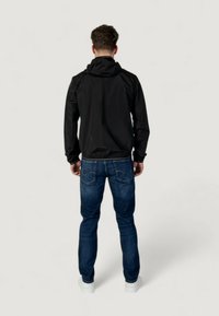 Giacca leggera nera con cappuccio e polsini elasticizzati, abbinata a jeans blu scuro e sneakers bianche, vista da dietro.