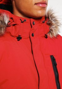 Parka rouge avec capuche en fourrure. Comprend des accents noirs, des boutons et une poche zippée à l'avant. Tissu texturé.