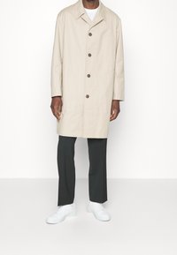 Beżowy płaszcz trench z kołnierzem i guzikami z przodu, zestawiony z ciemnymi spodniami i białymi sneakersami. Płaszcz ma czysty, minimalistyczny design.