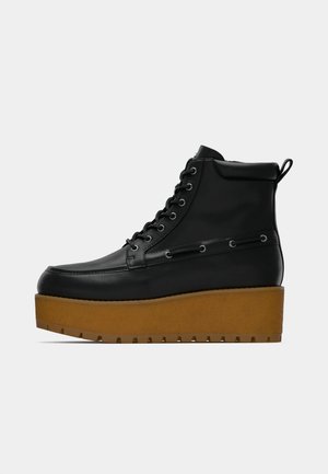 Botte montante en cuir noir avec semelle plateforme épaisse marron et fermeture à lacets, sur un fond gris uni.