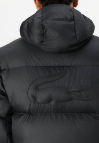 Doudoune noire avec capuche, présentant une finition texturée et un logo de crocodile brodé dans le dos.