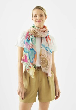 Vrouw draagt een wit T-shirt, beige korte broek met riem en een kleurrijke sjaal met abstracte en door de natuur geïnspireerde patronen.