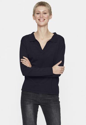 Navy long-sleeve polo shirt with a V-neck, made from textured fabric. Model with short hair stands with arms crossed, wearing dark jeans.