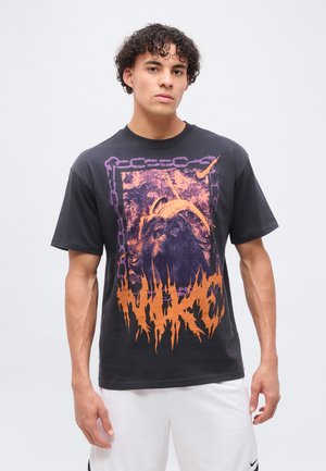 Jeune homme portant un t-shirt graphique noir avec un motif de tête de chèvre orange et violet et un short blanc Nike, debout sur un fond uni.