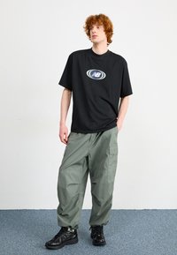 Czarny t-shirt z krótkim rękawem z białym i niebieskim logo, w połączeniu z luźnymi oliwkowymi spodniami cargo oraz czarnymi butami sportowymi.