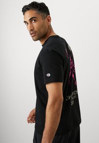 Schwarzes T-Shirt aus Baumwolle, mit einem bunten Grafikdesign auf der Rückseite und einem kleinen Logo-Patch auf dem linken Ärmel. Kurze Ärmel.