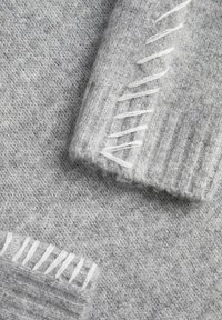 Tissu de pull en maille gris avec des poignets côtelés, présentant des accents de couture blanche dans un motif droit. Texture douce, avec une minimalité de duveteux visible.