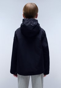 Giacca nera con cappuccio, rifinitura elasticizzata, tessuto morbido e vestibilità comoda. Vista da dietro, indossata sopra pantaloni della tuta grigi.