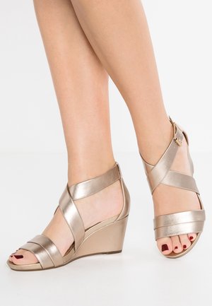 Pieds avec un vernis à ongles rouge foncé portant des sandales compensées dorées métalliques avec des brides croisées et une boucle à la cheville sur fond blanc.
